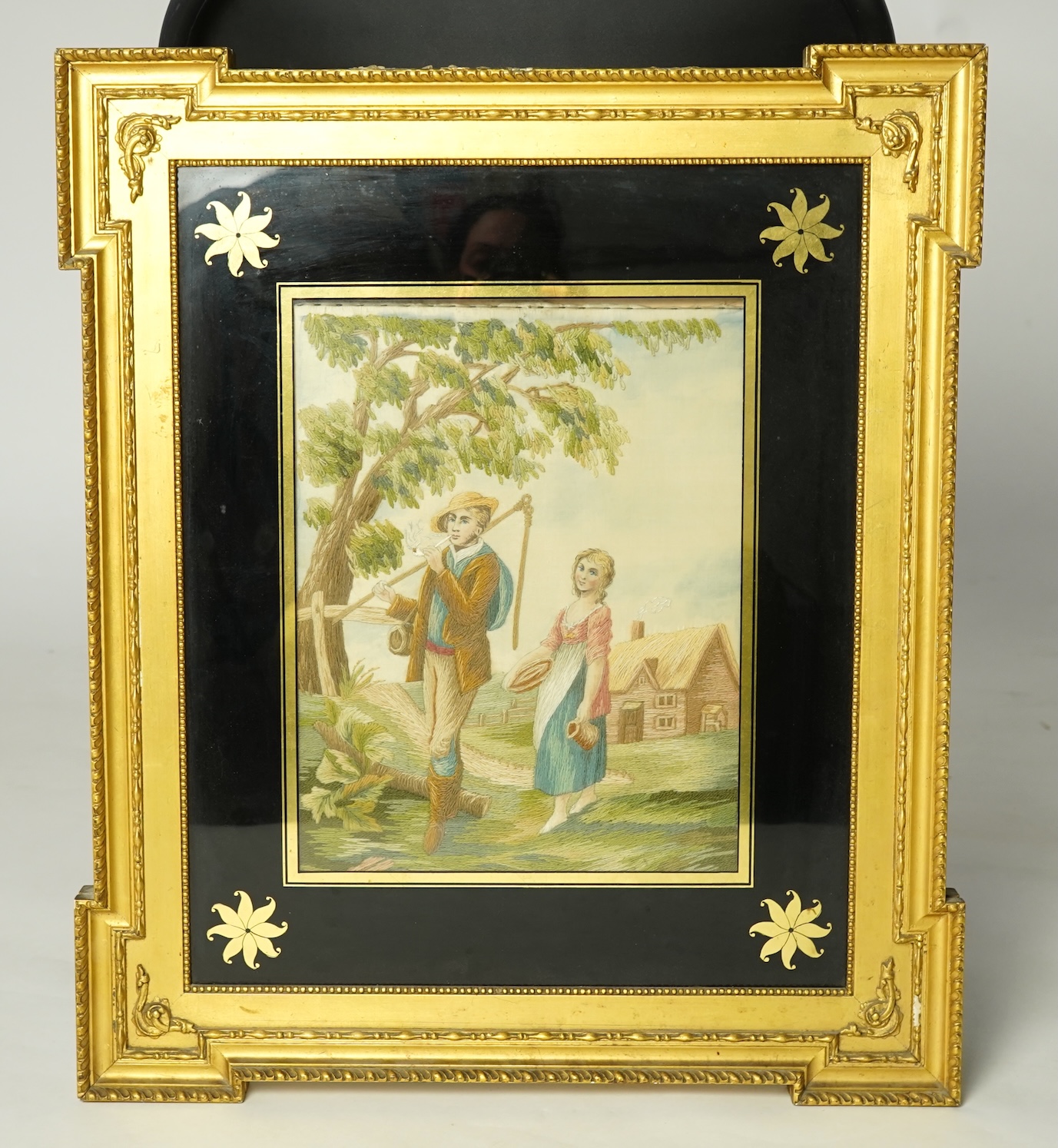An early 20th silk embroidery of a boy and a girl in a country landscape by a tree and cottage. Worked in stem stitch with painted faces hands and background. Mounted with Verre Eglomise glass and a gilt rectangular fram
